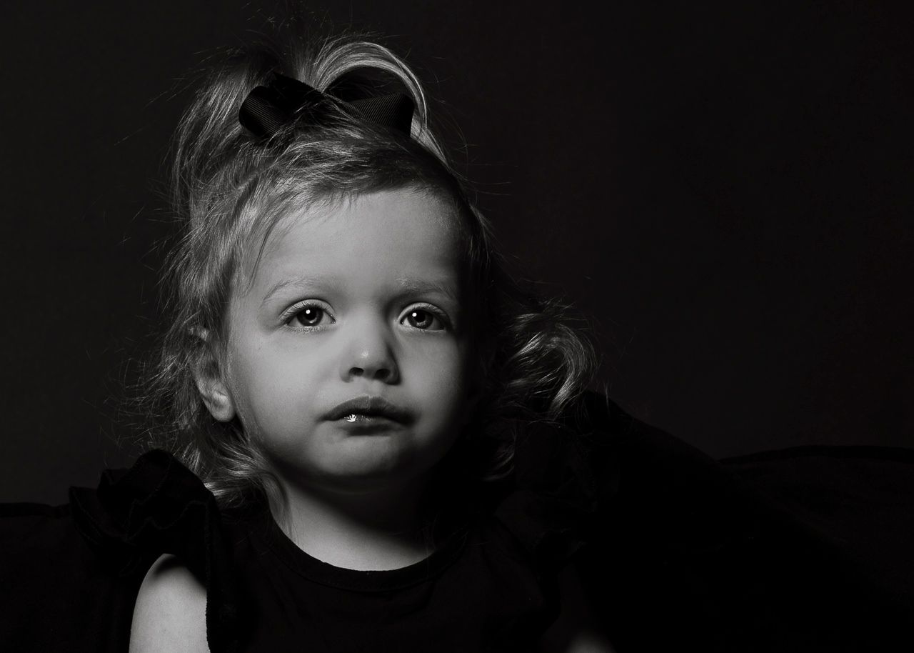 Black and white portrait of a young child with light hair wearing dark clothing against a dark background.