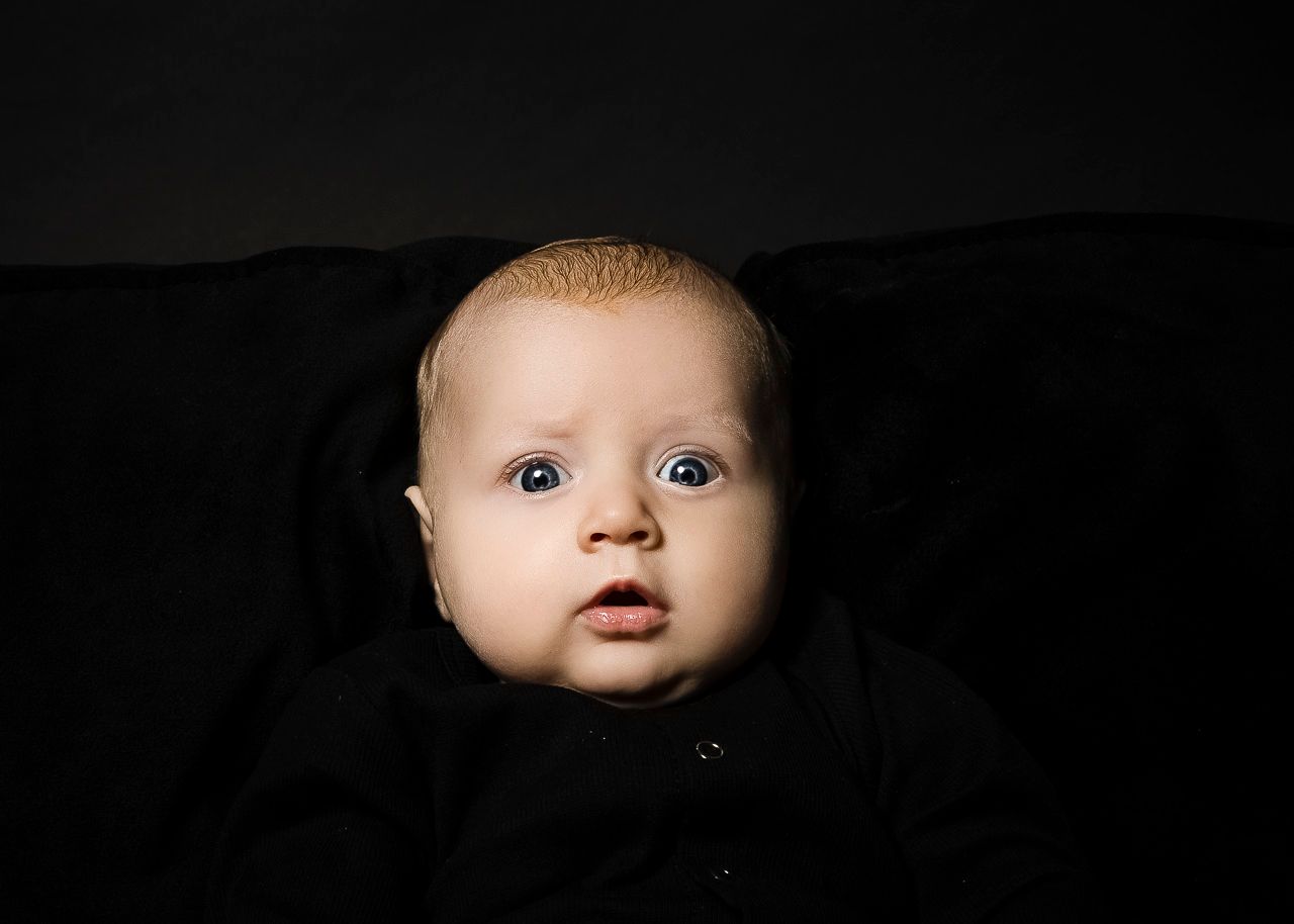 Baby wearing black clothing is photographed against a black background with dramatic lighting effect.