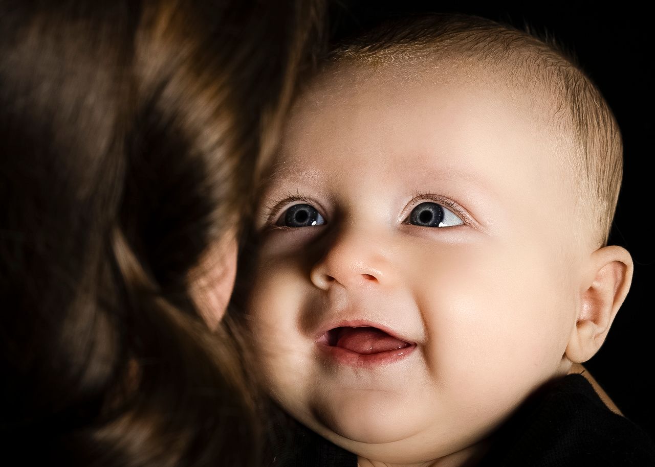 A smiling baby with bright eyes looks up at mother, captured in warm lighting against dark background.