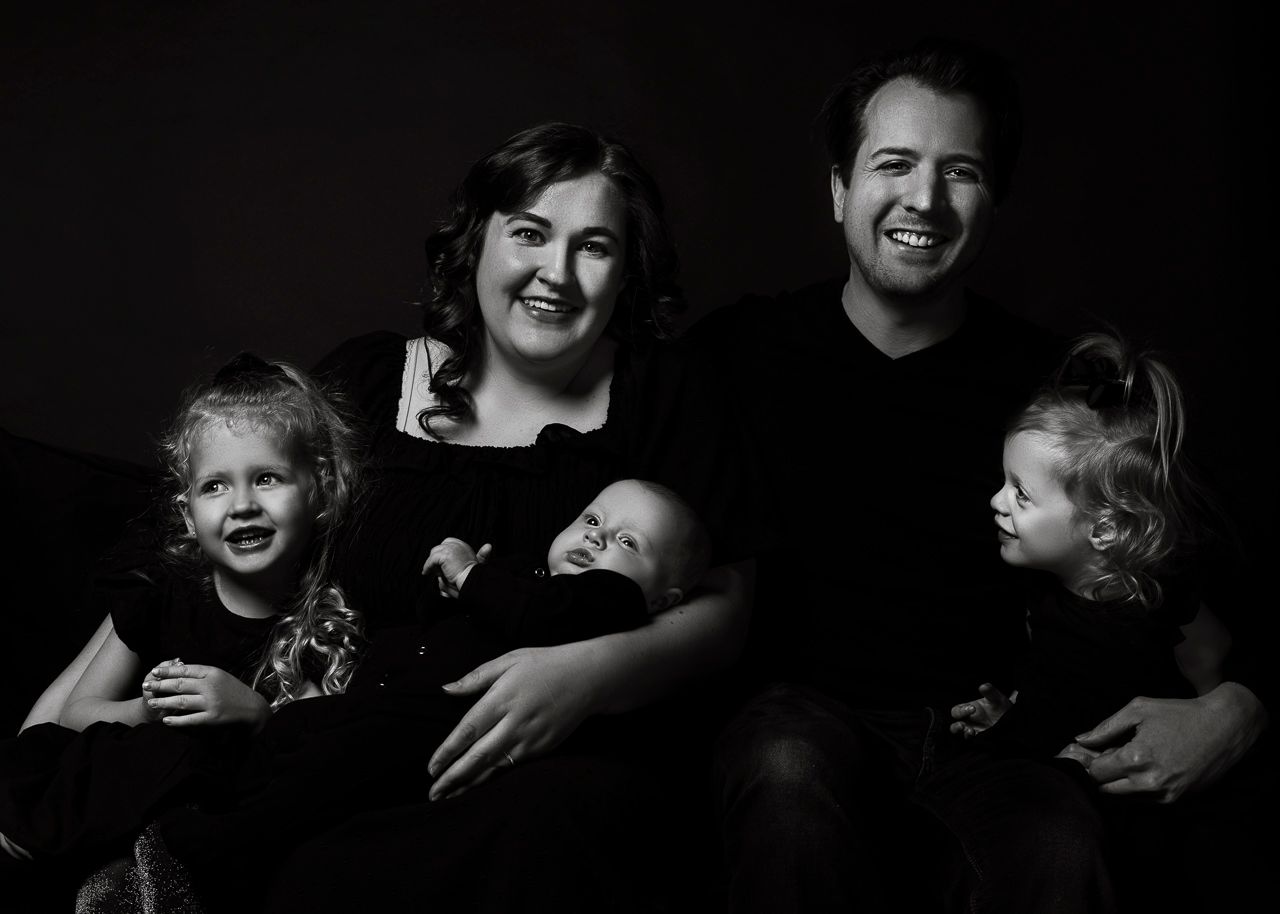 Black and white studio portrait of a family of five sitting close together against a dark background.