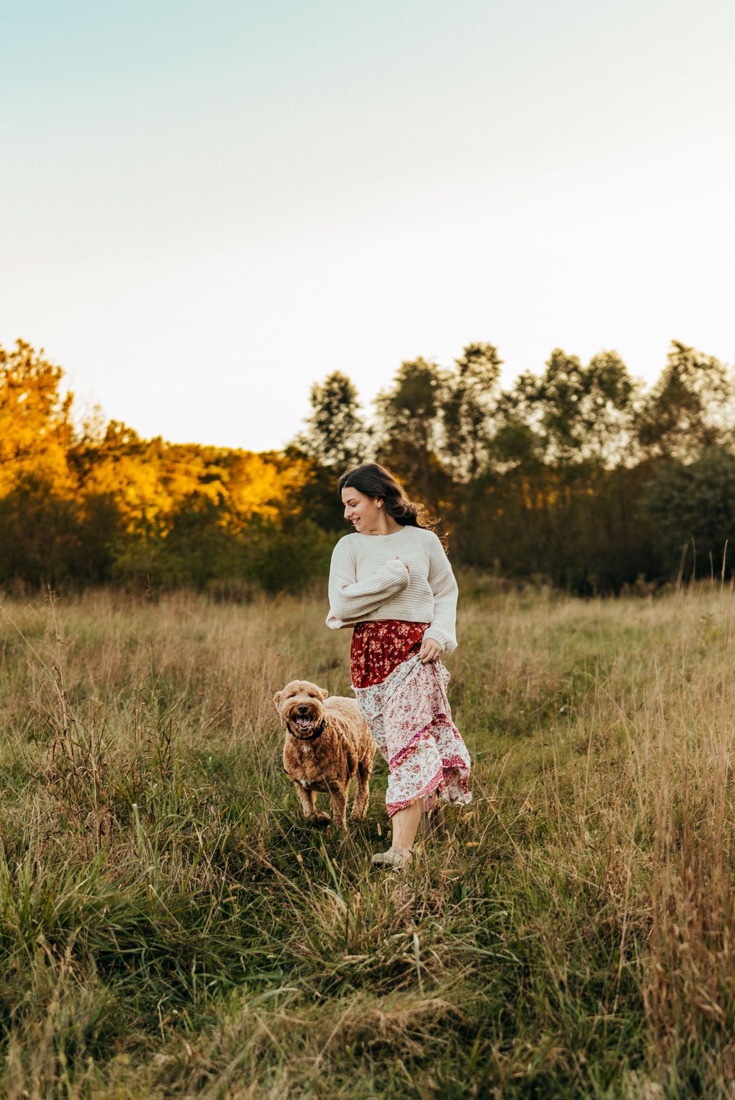 Person in flowing skirt walking with dog through tall grass at sunset in poplar grove IL