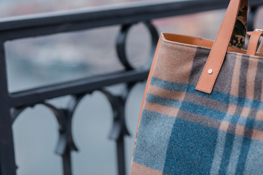 Closeup of a plaid tote bag in beige and blue tones with leather handles against a metal railing.