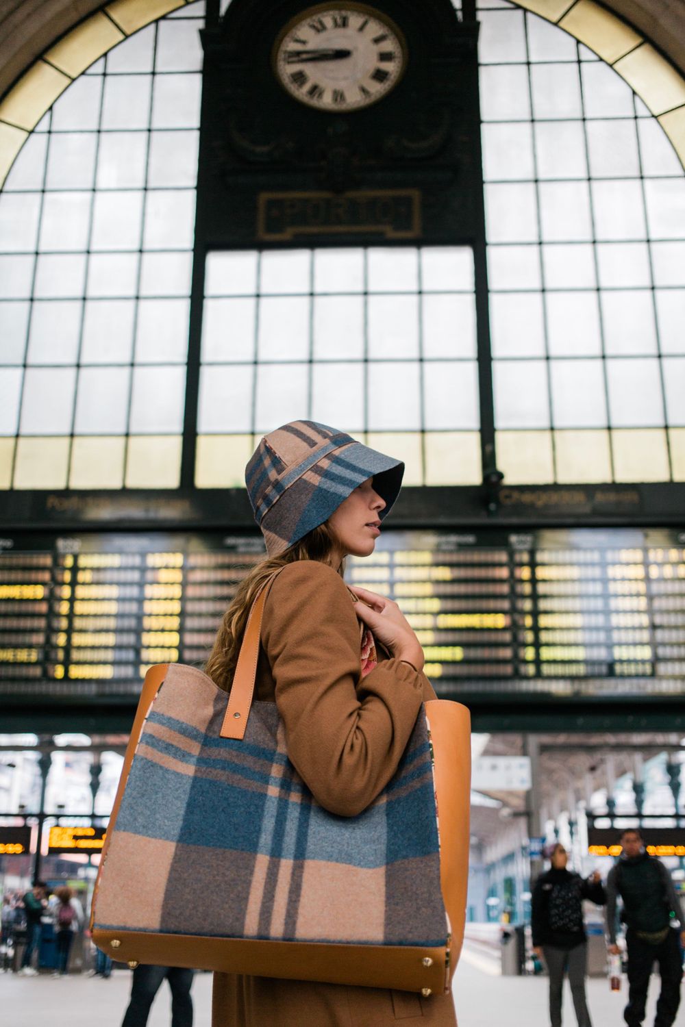 Stylish traveler with plaid hat and matching bag stands beneath vintage train station clock at busy terminal.