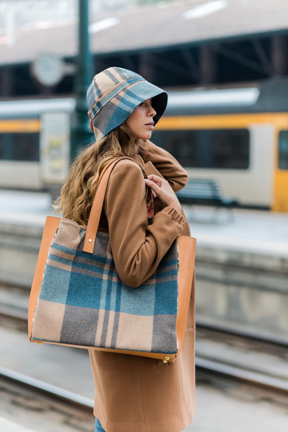 Stylish winter outfit featuring a plaid cap and matching tote bag with a camel coat at a train station platform.