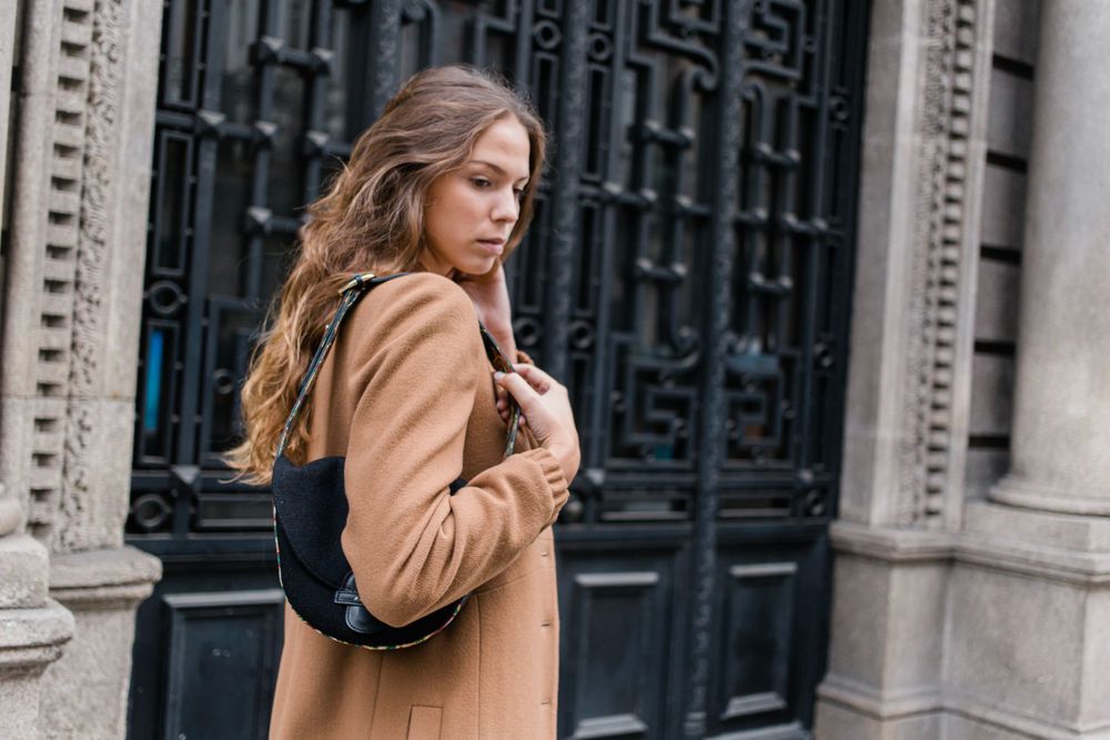 A woman in a tan coat walking past ornate dark doors of a historic building with architectural details.