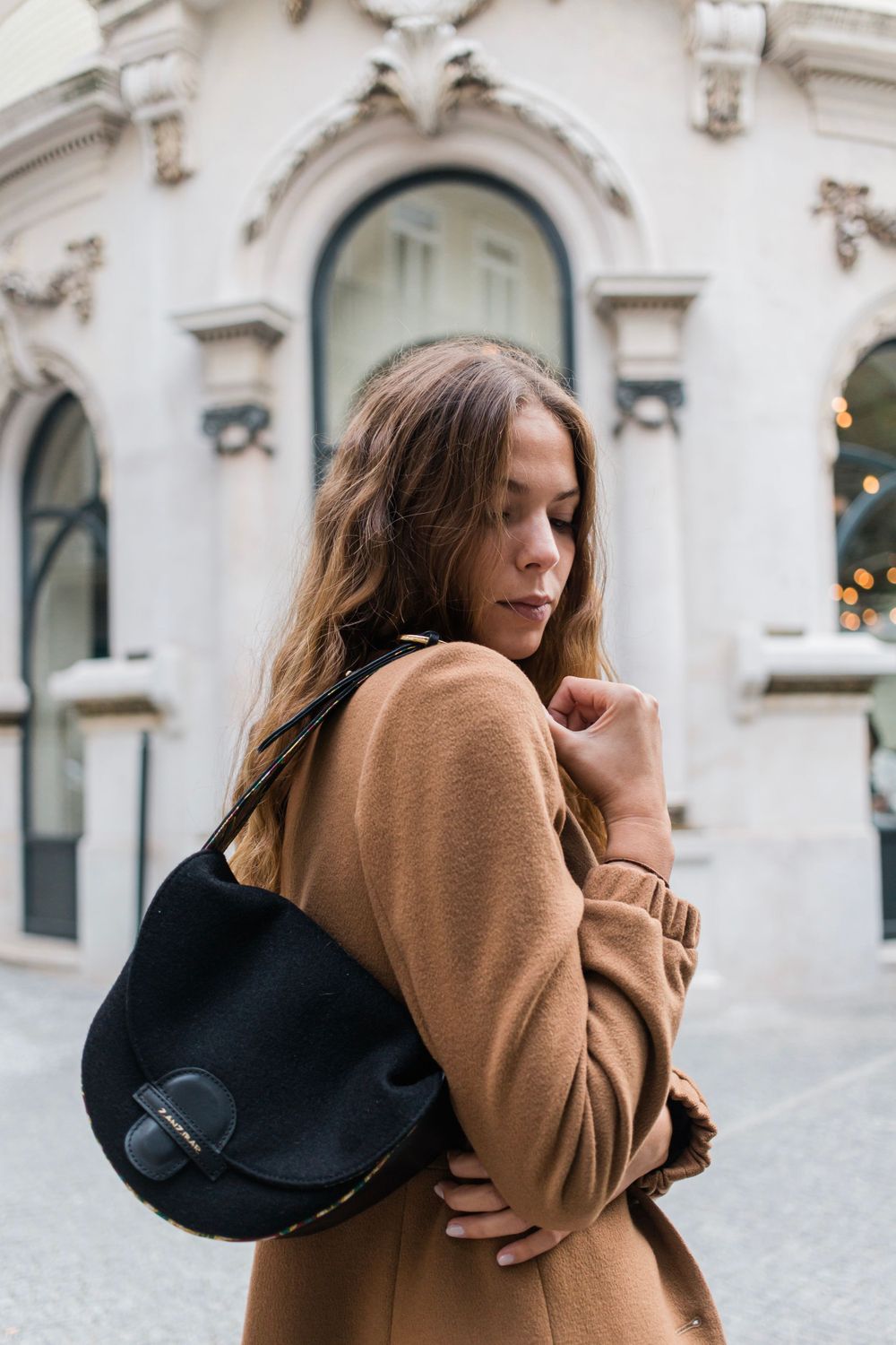 A person wearing a camel coat and black leather bag poses on a street with ornate architecture in the background.