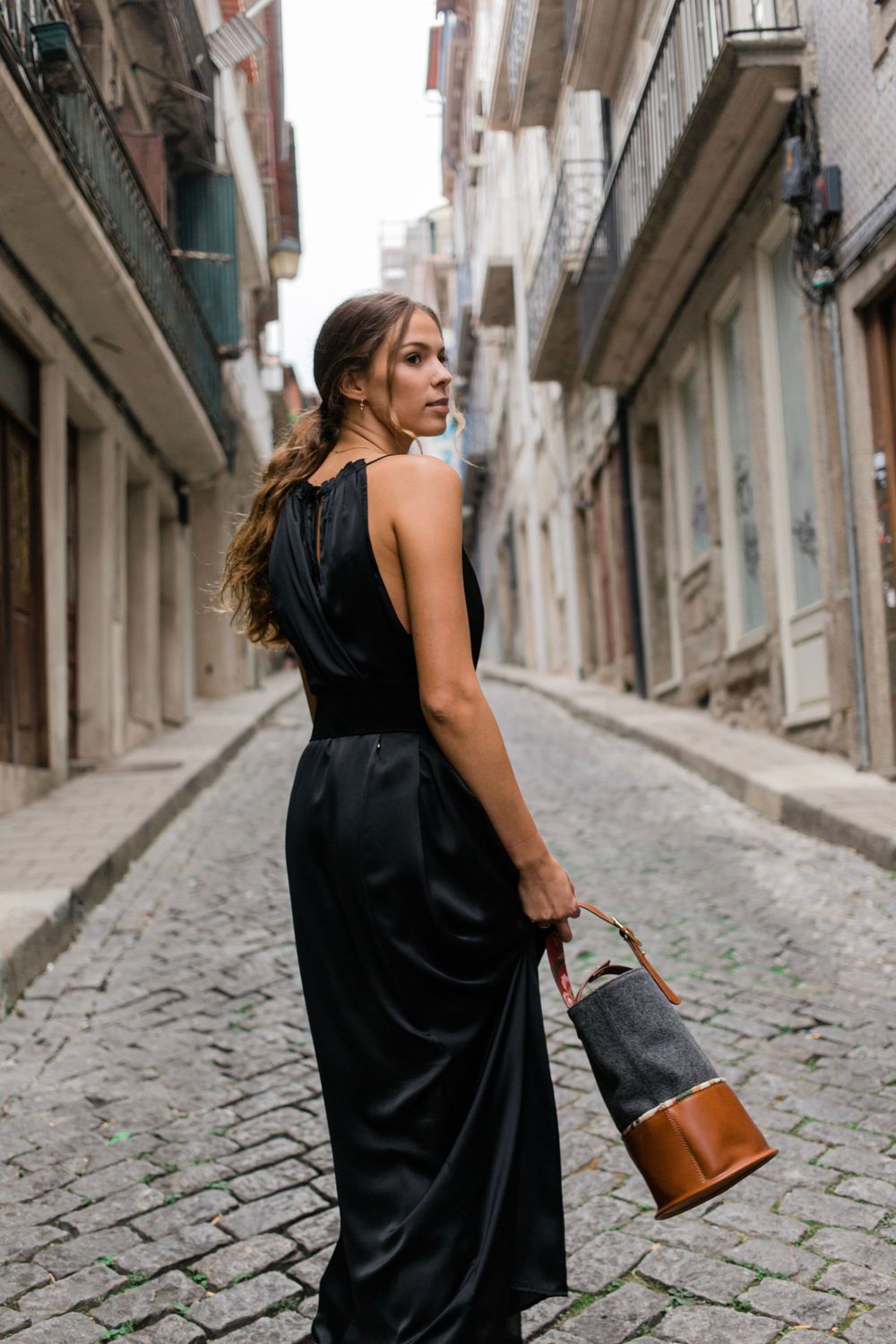 Woman wearing elegant black dress stands in narrow cobblestone alley carrying leather handbag in European city.