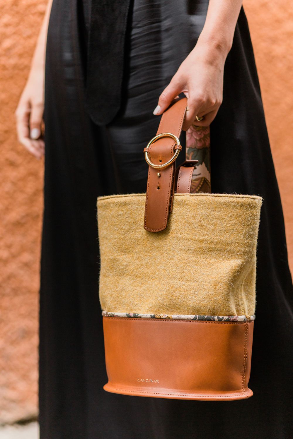 Hand holding stylish two-tone bucket bag with tan leather bottom and beige canvas top against terracotta wall background.
