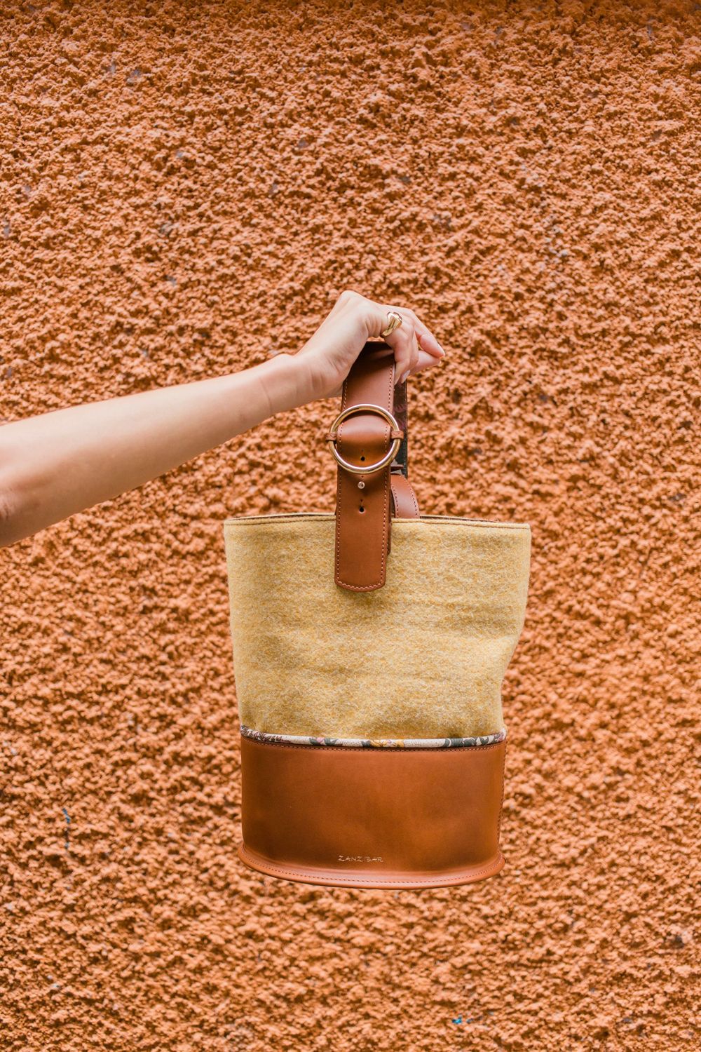 Hand holds a stylish two-tone bucket bag with canvas and leather against a textured terracotta wall.