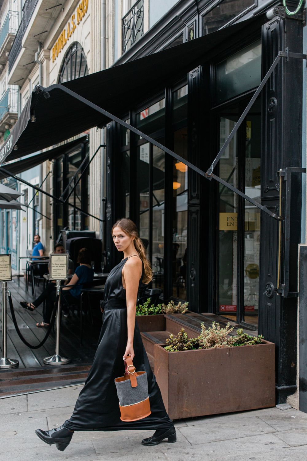 Someone in a long black dress and boots walks past a storefront with black awnings and planters on a city street.