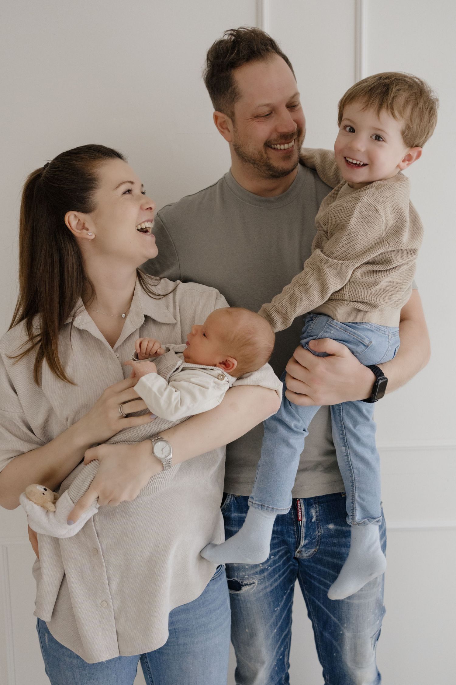 A family shares joyful laughter together while holding their newborn baby and toddler against a white wall.
