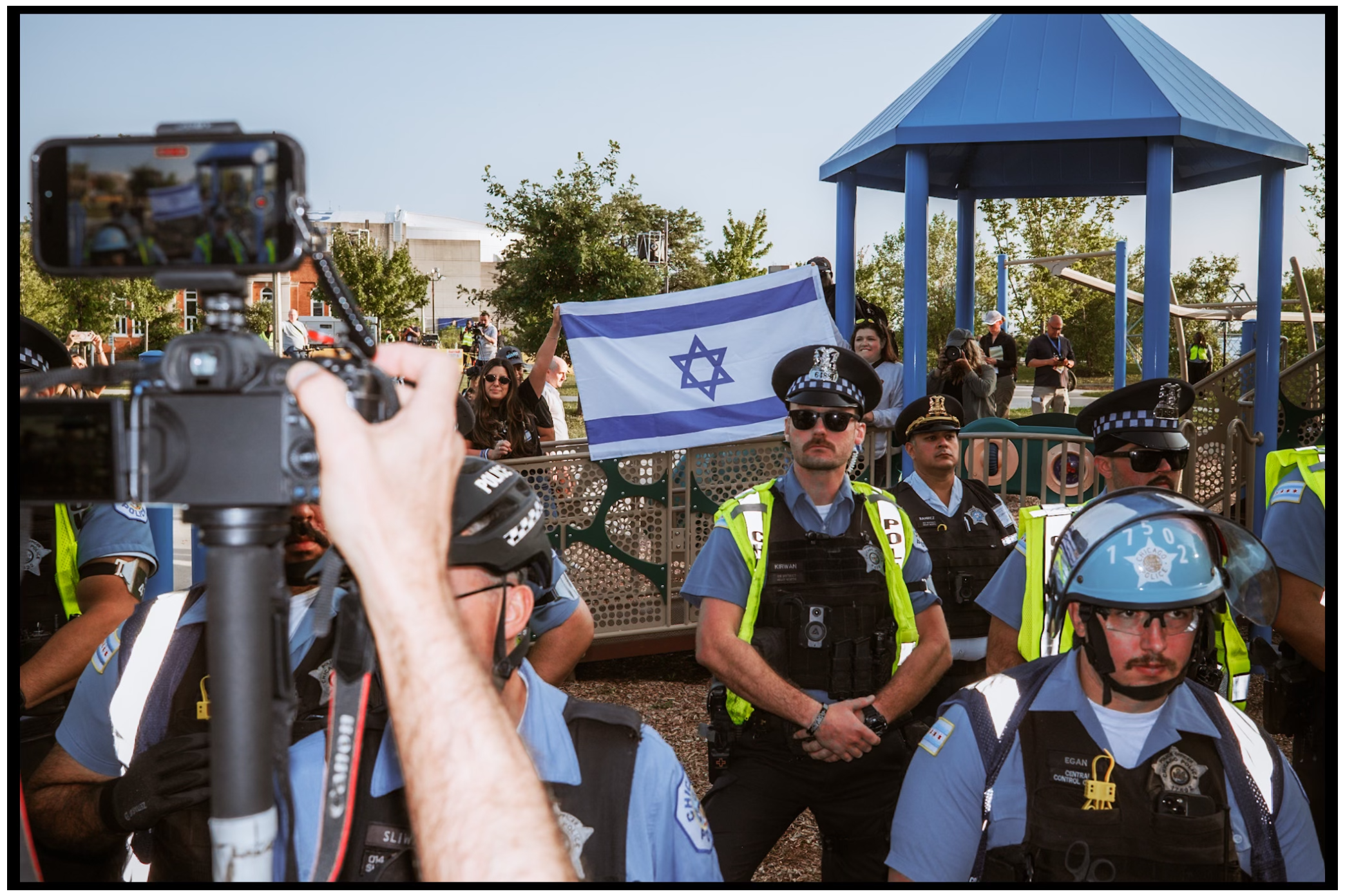 Police officers in blue uniforms stand guard as someone holds phone with Israeli flag in background. Chicago. March on DNC