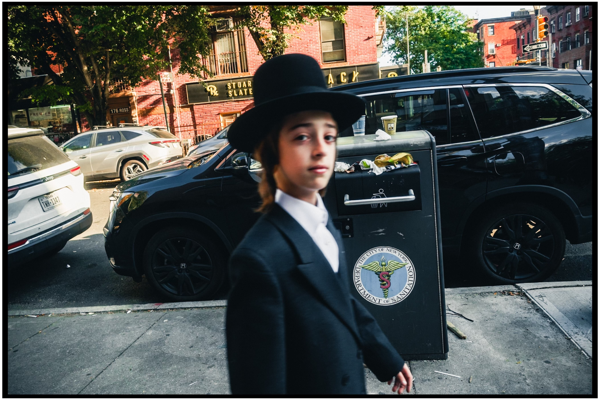 A person in traditional Orthodox Jewish attire including a black hat stands on a city street near parked cars. Kapparot, Brooklyn NYC