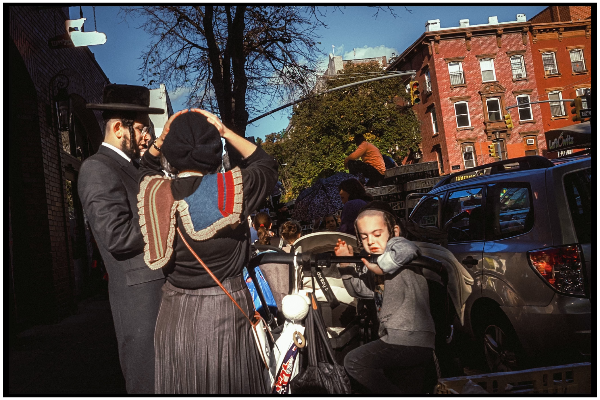 Several people gathering around a stroller on a city street with brick buildings in the background. Kapparot. Brooklyn.