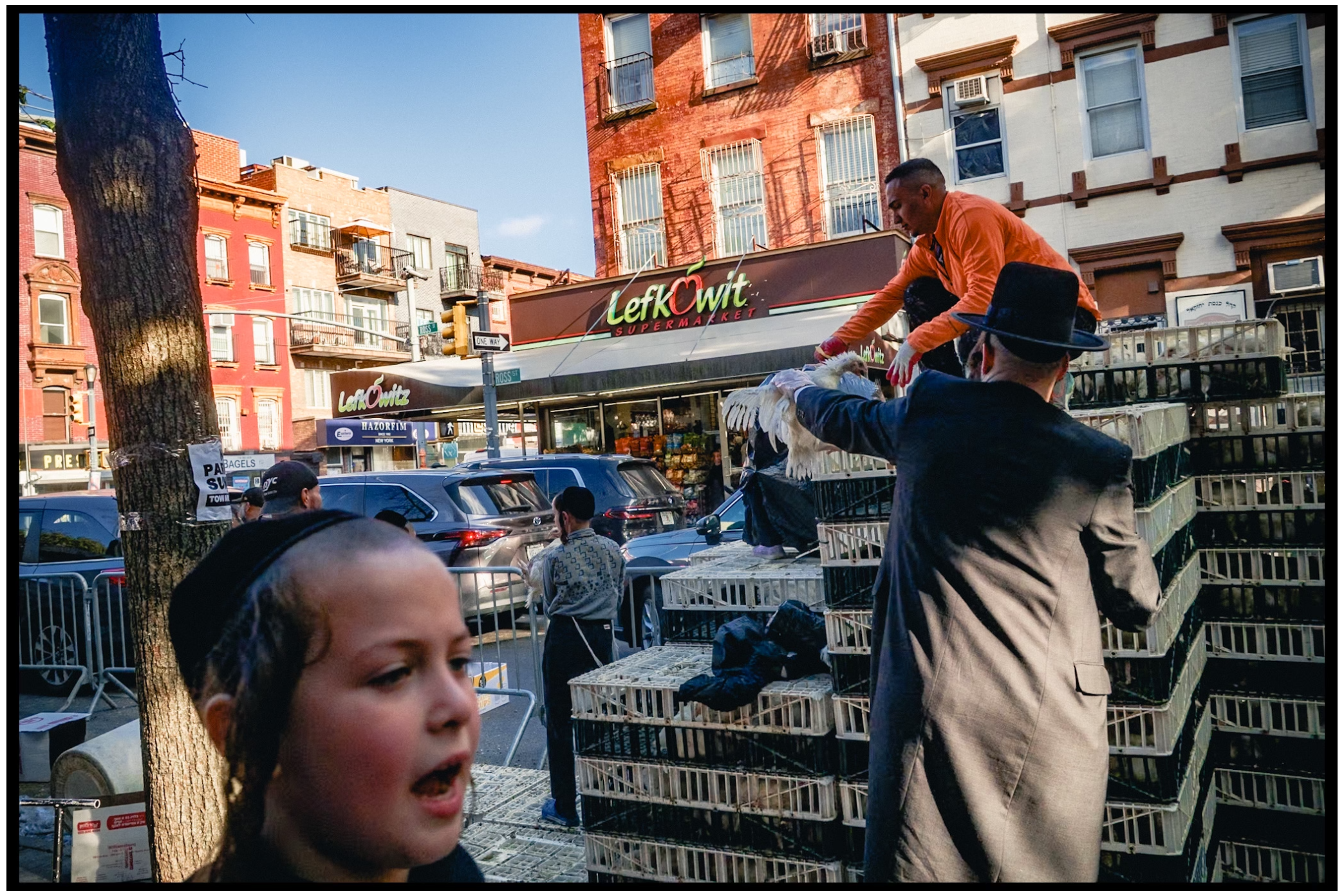 Dynamic street scene with people interacting around stacked boxes in front of brick buildings. Kapparot, Brooklyn NYC