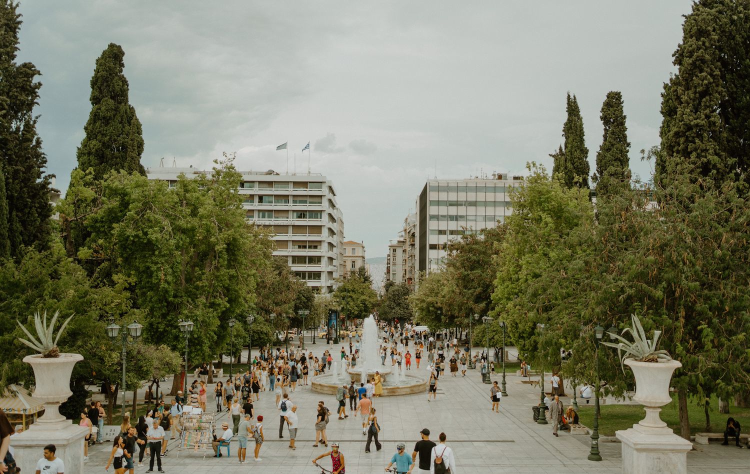 People walking along tree-lined Syntagma Square with modern buildings and fountains in Athens, Greece.