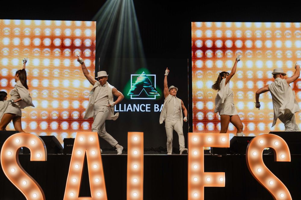 Dancers perform in white costumes against an illuminated backdrop with SALES text.