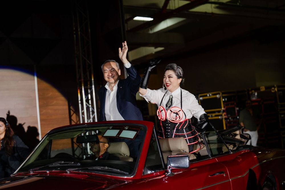 Two people wave while standing in a vintage red convertible car during an indoor event.