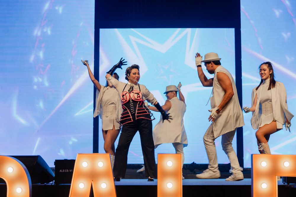 Dancers perform on stage with illuminated letters and blue backdrop screens.
