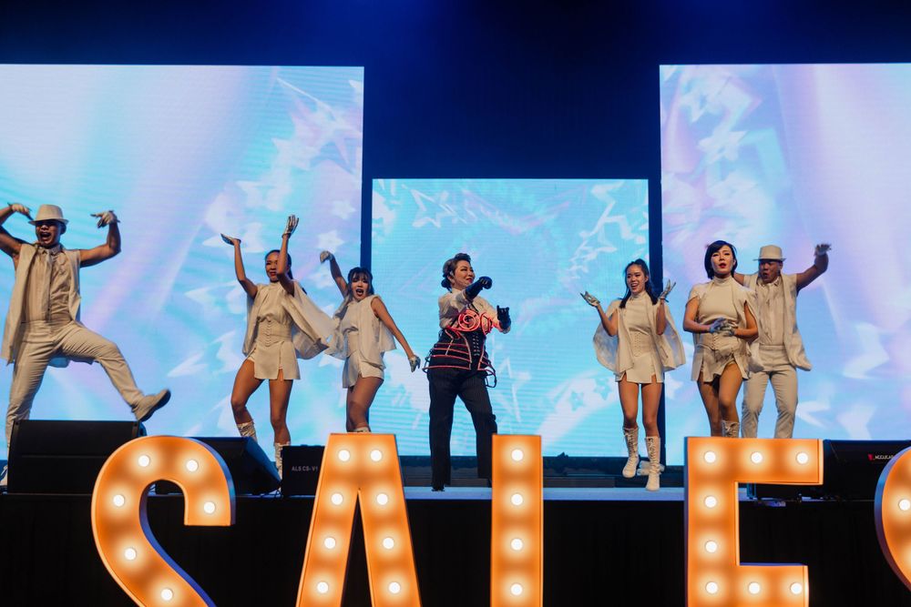 Performers dancing on stage with large illuminated SALES letters and video screens in background.