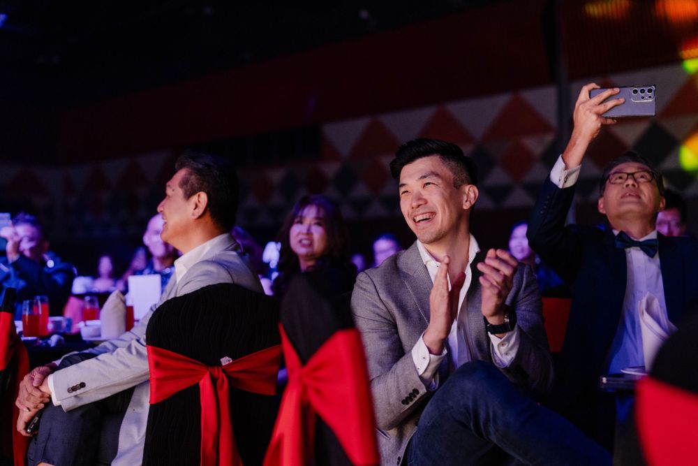 Audience members in formal attire watching and applauding at an event with red seat decorations.