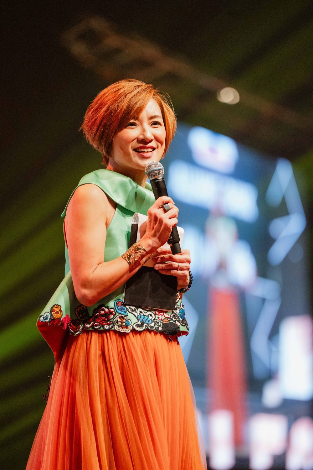 Performer in orange skirt and green top singing on stage with dramatic lighting.