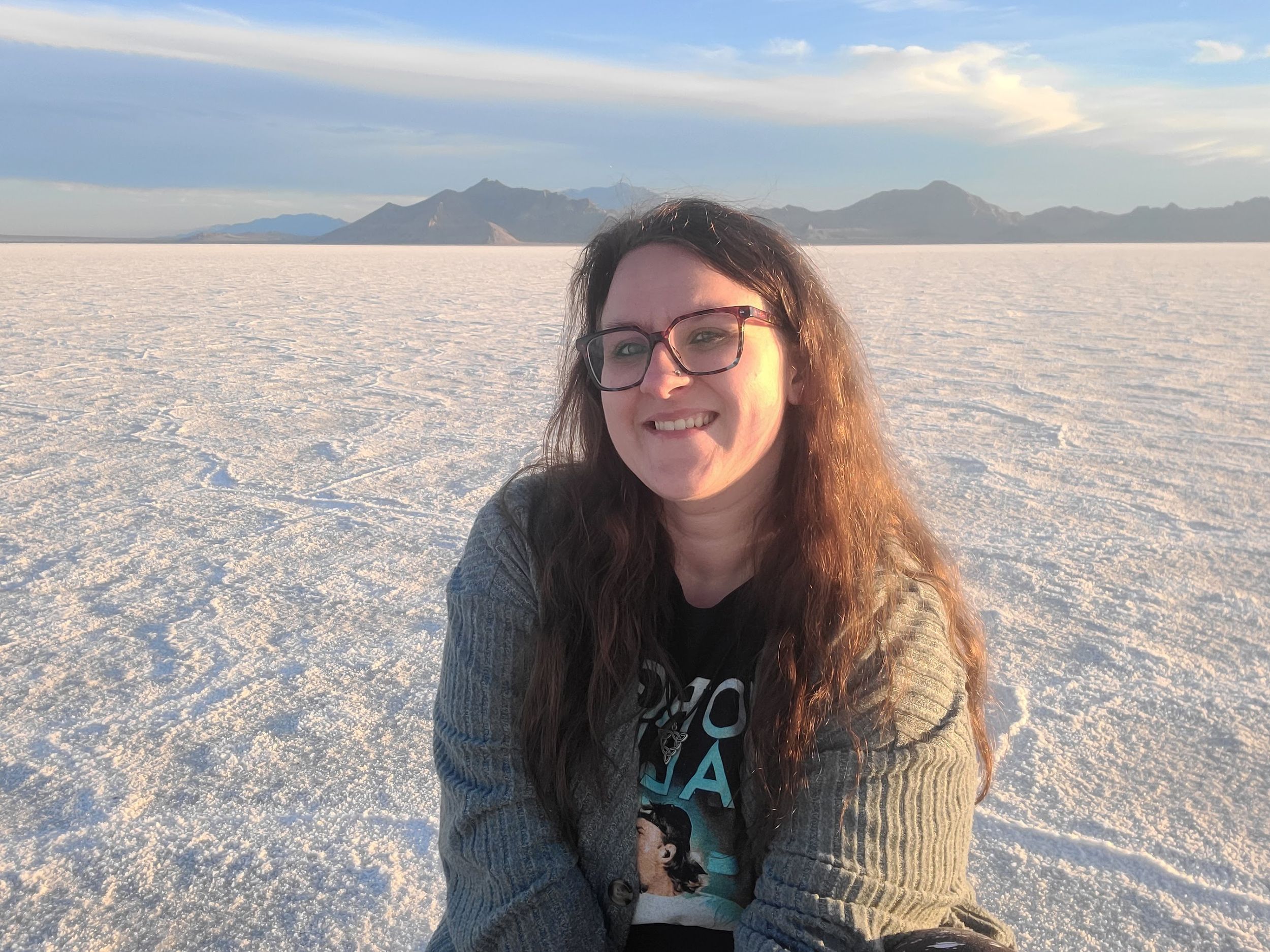 Someone smiles while sitting on the bright white salt flats with mountain ranges visible in the distance against blue skies.
