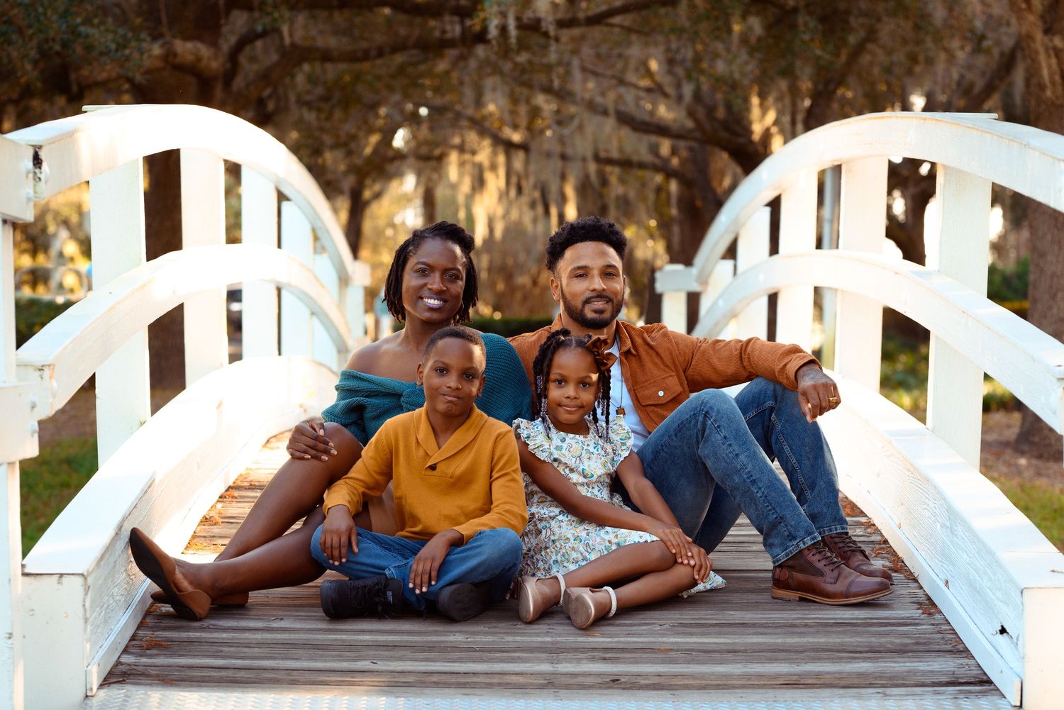 A family sits together laughing and smiling on a white wooden bridge in a candid outdoor portrait.