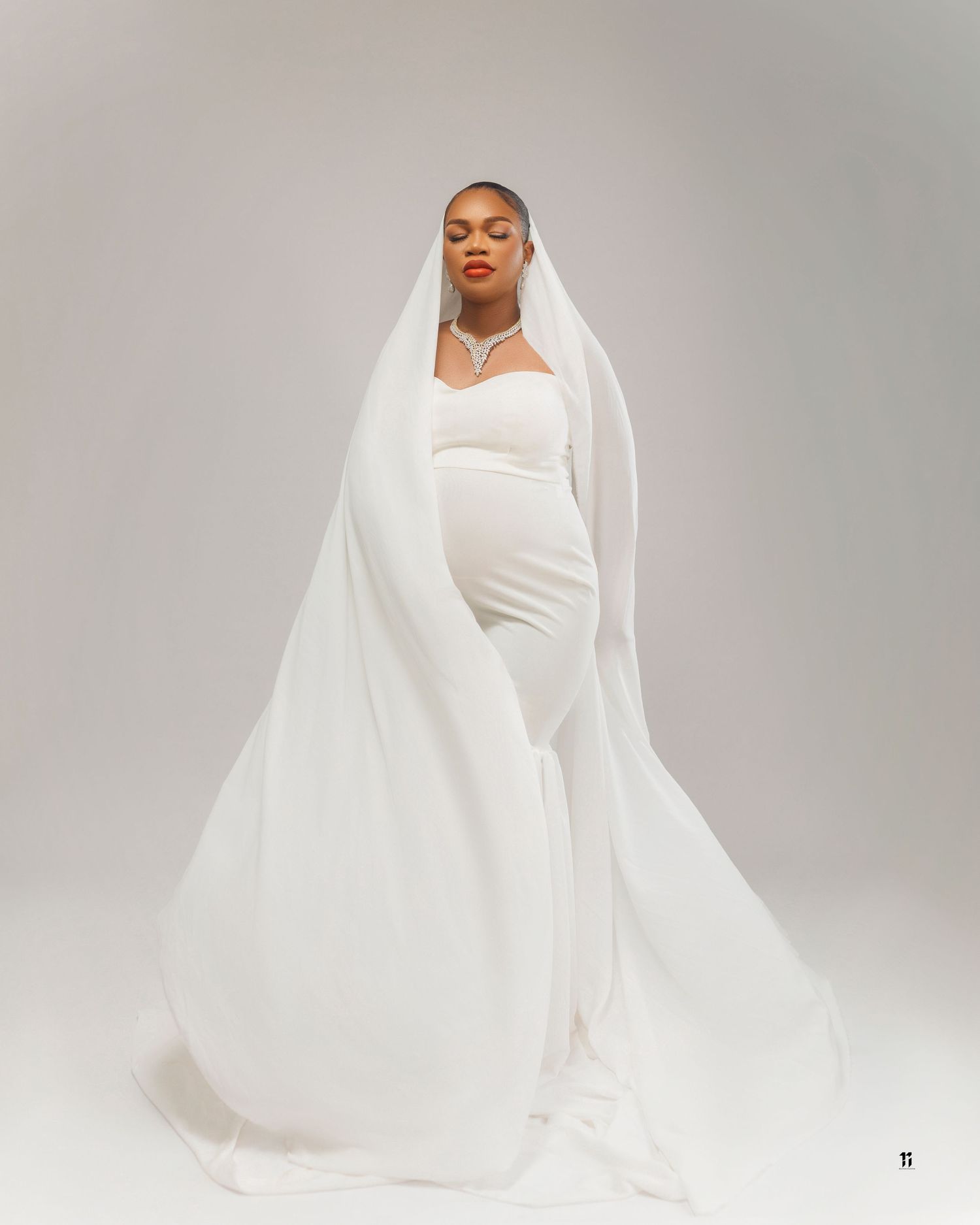 A bride in an elegant white form-fitting wedding dress and flowing veil poses against a light gray backdrop.