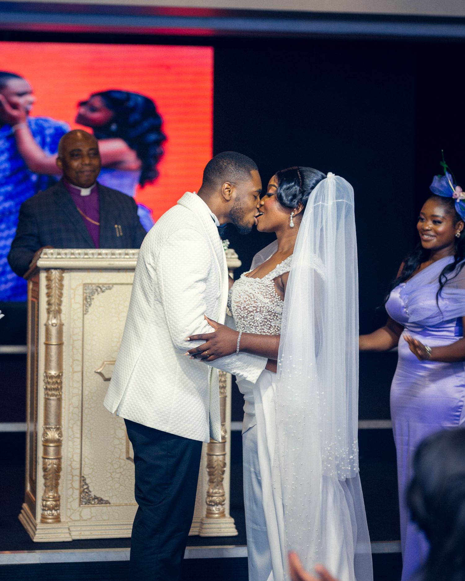 Newlyweds share their first kiss at the altar with elegant decor and wedding party looking on.