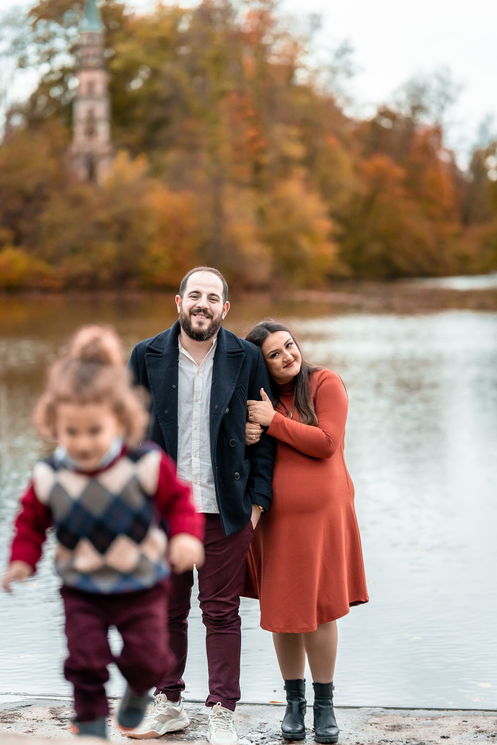 Family moment by autumn lake with child in foreground and expecting parents.