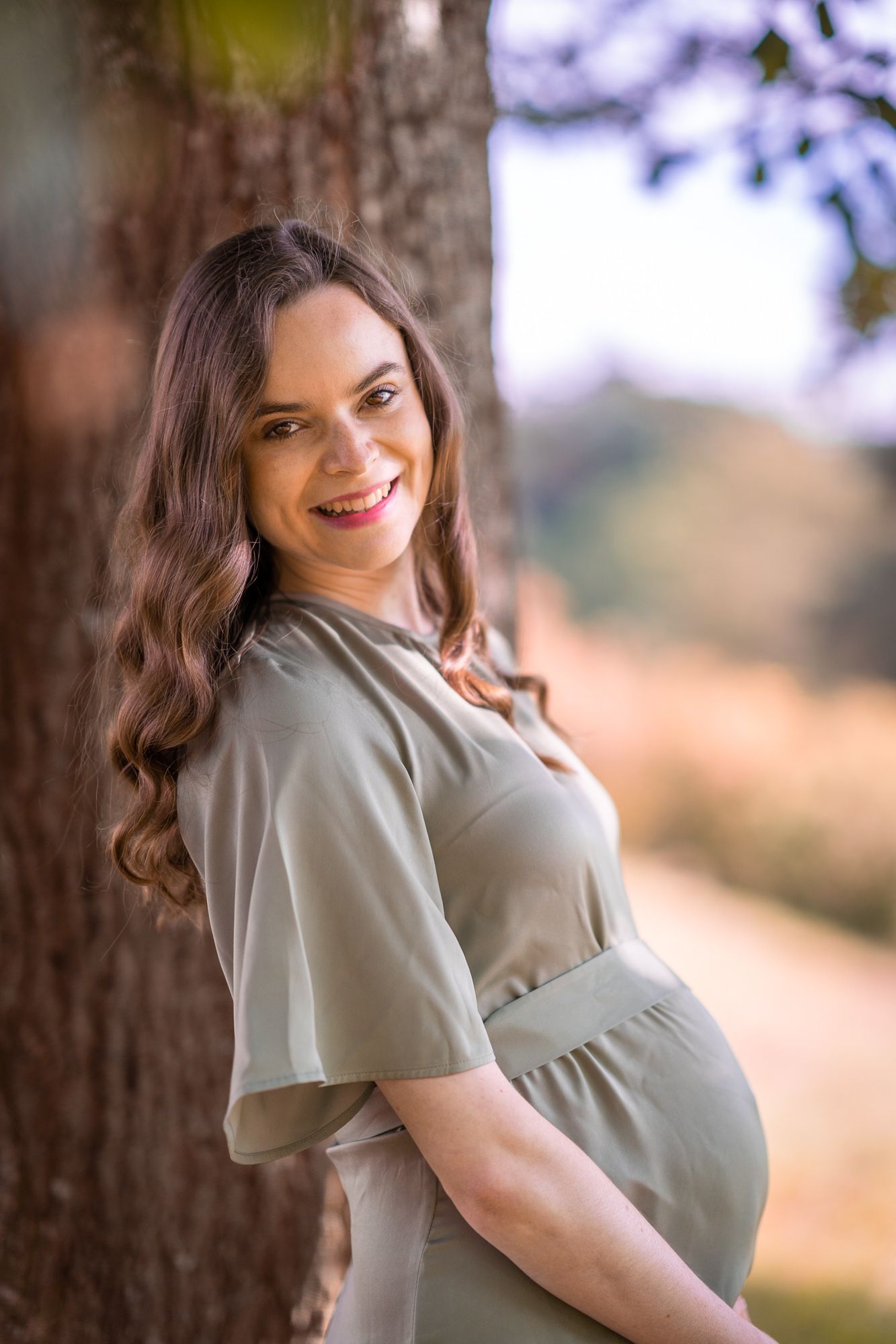 Maternity portrait of woman in light dress leaning against tree in natural setting.