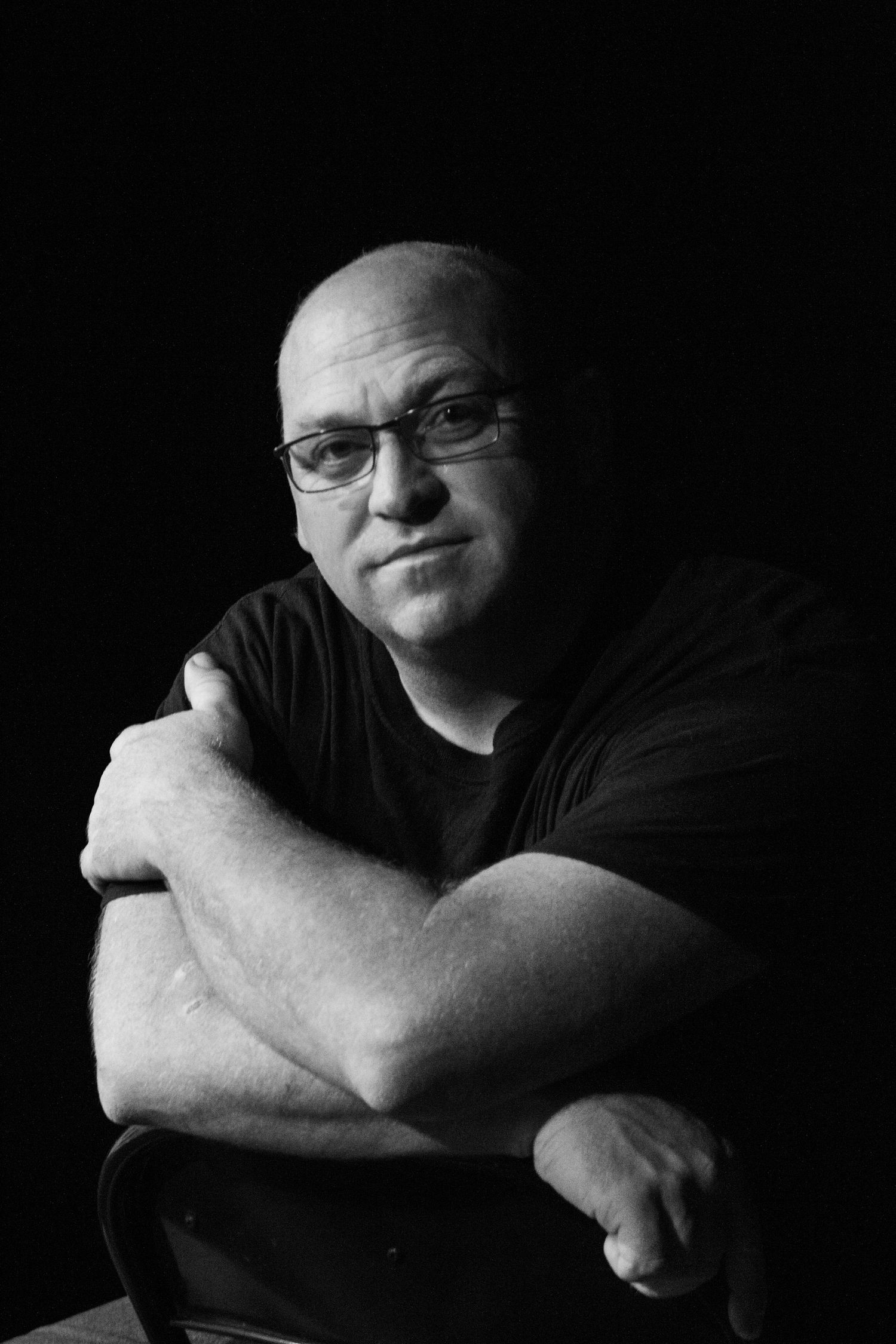 Black and white portrait of someone wearing glasses and a dark t-shirt with crossed arms looking at camera.