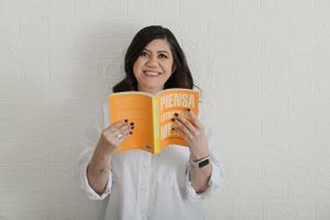 A person in a white button-down shirt smiles while holding an orange book against a light gray wall.