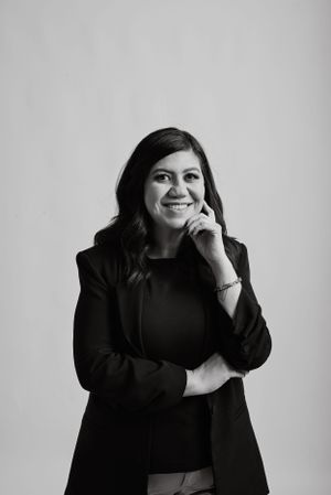 Professional black and white portrait of someone in a dark blazer smiling with hand thoughtfully posed near chin.