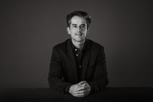 Professional black and white portrait of a man in a dark blazer seated at a table against a dark background.