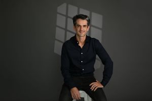 Professional studio portrait of a person in a dark button-down shirt seated against a gray background with dramatic lighting.