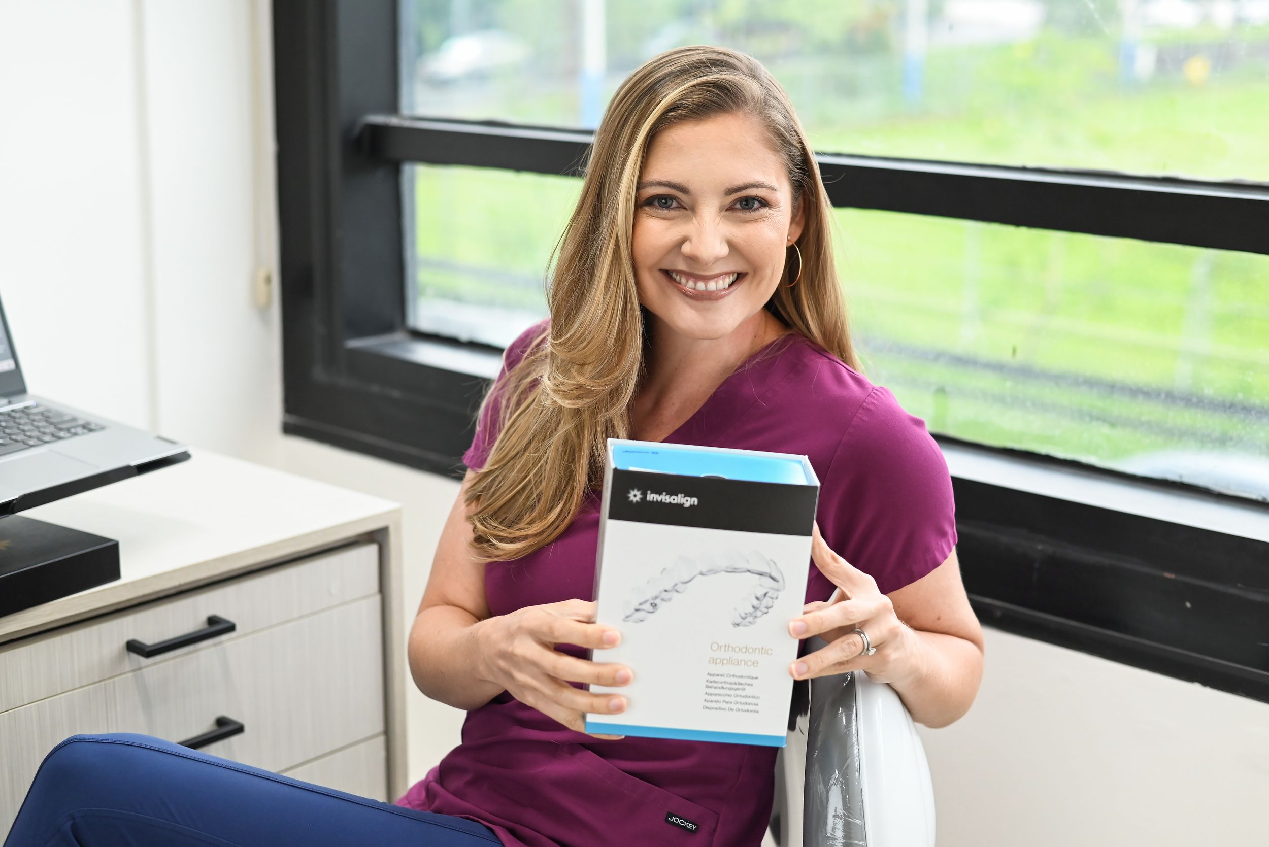 Healthcare professional in burgundy scrubs holds up a medical folder in a bright office setting near a window.
