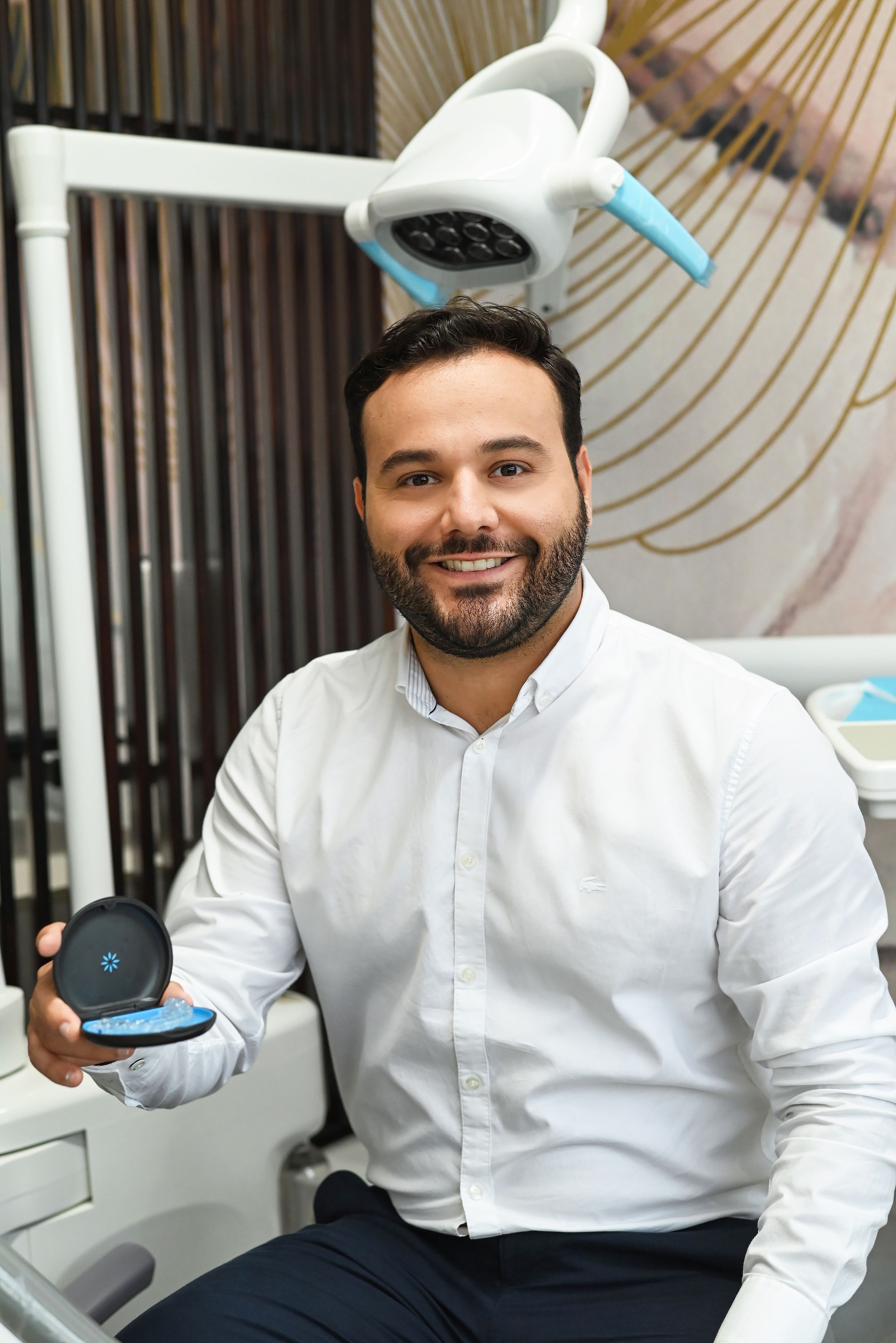 Dental professional in white shirt poses at workstation with dental equipment and examining light in modern office.