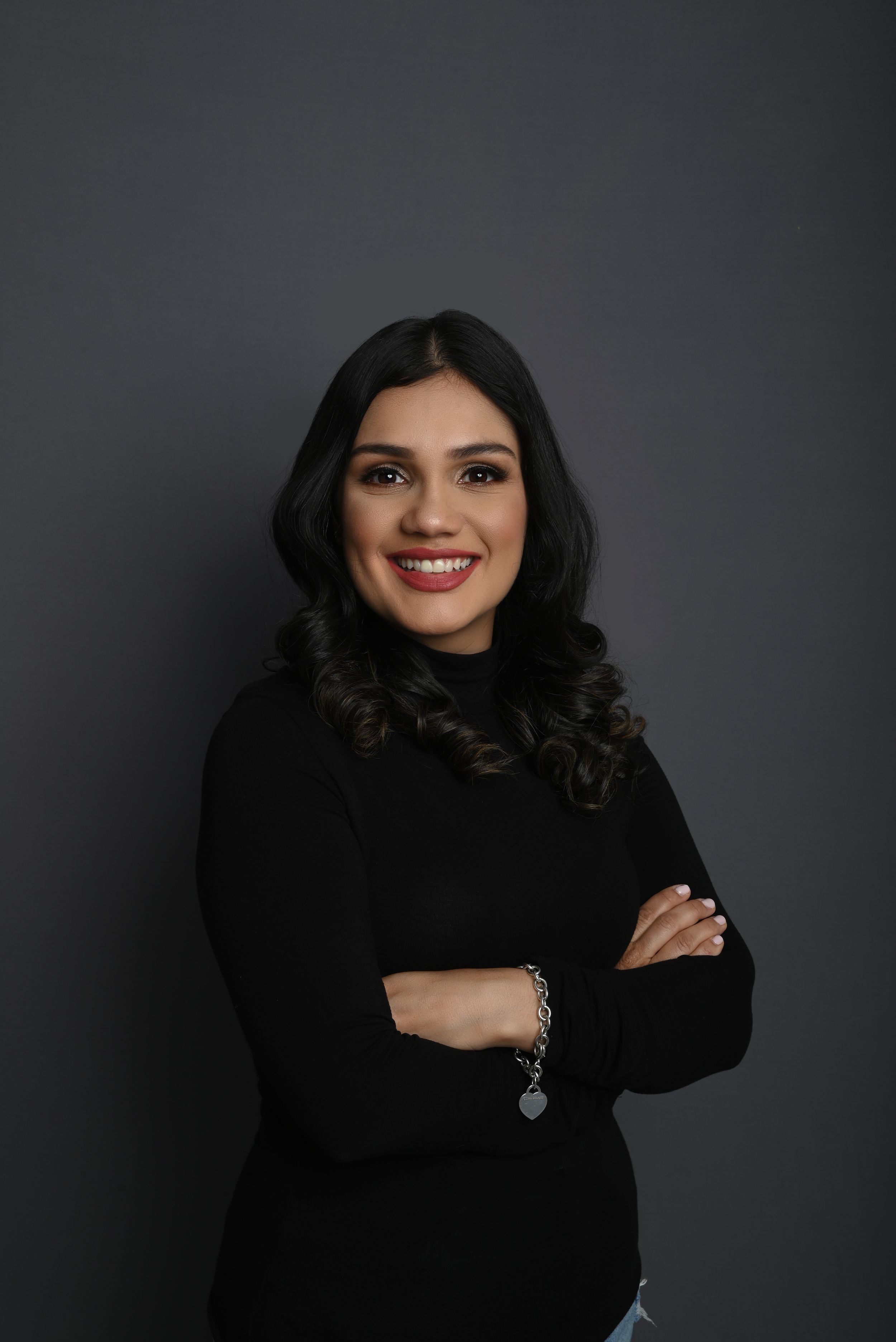 Portrait of woman in black sweater with fur collar smiling against dark gray background.