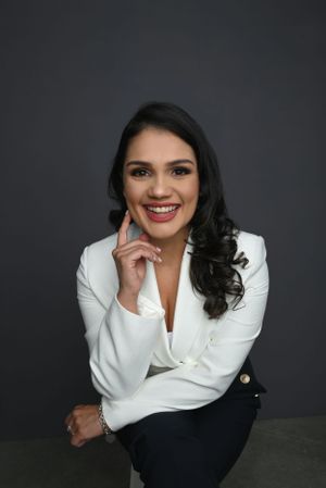 Professional portrait of someone in a white blazer and dark pants sitting casually against dark background with a warm smile.