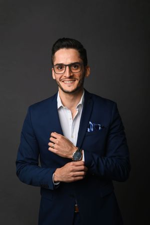 Professional portrait of someone in a navy blue suit and glasses against a dark background, smiling warmly.