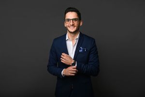 A professionally dressed person in a navy blazer and glasses smiles confidently against a dark background.