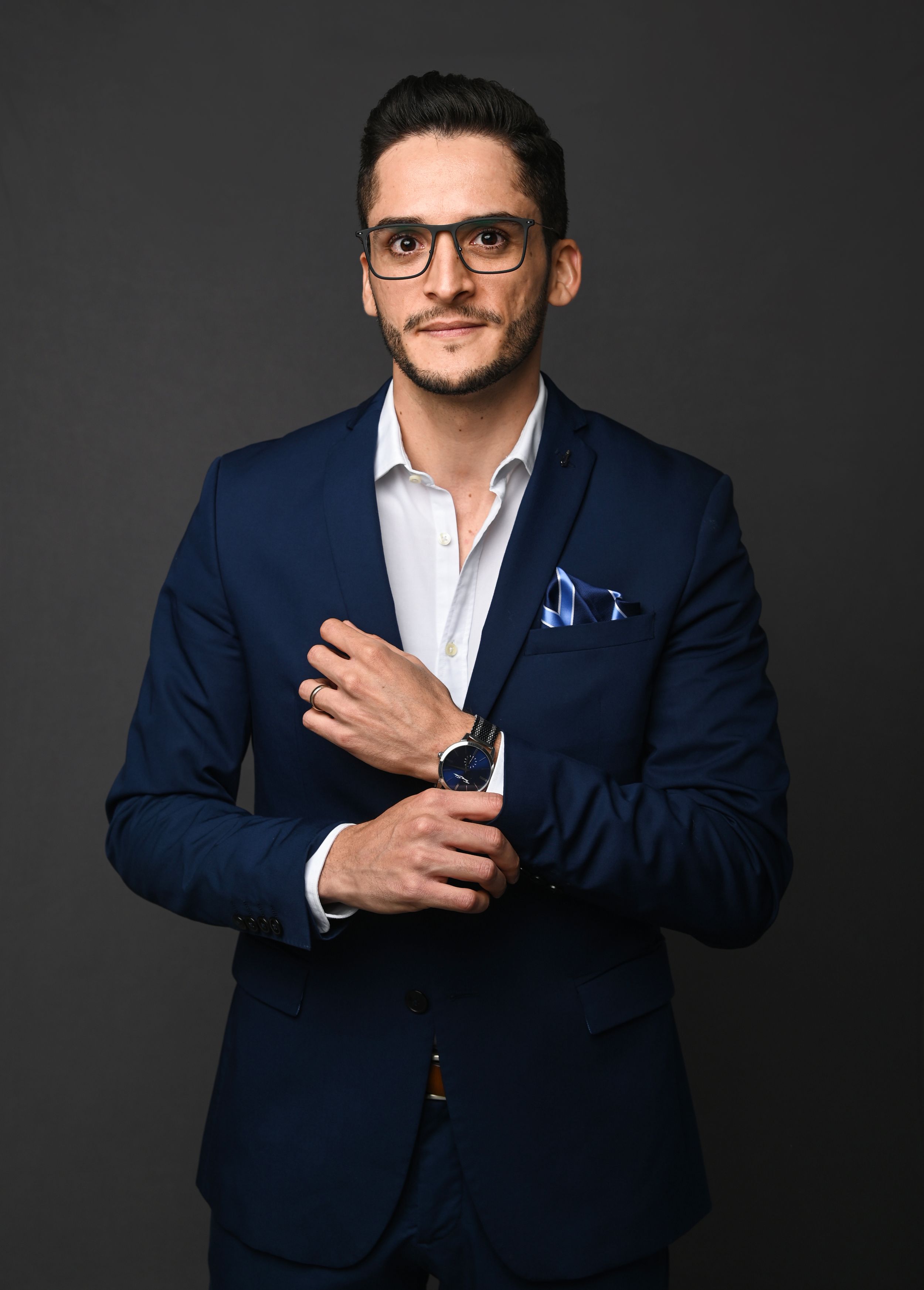 Professional portrait of someone in a navy blazer, white dress shirt, glasses and pocket square against dark background.