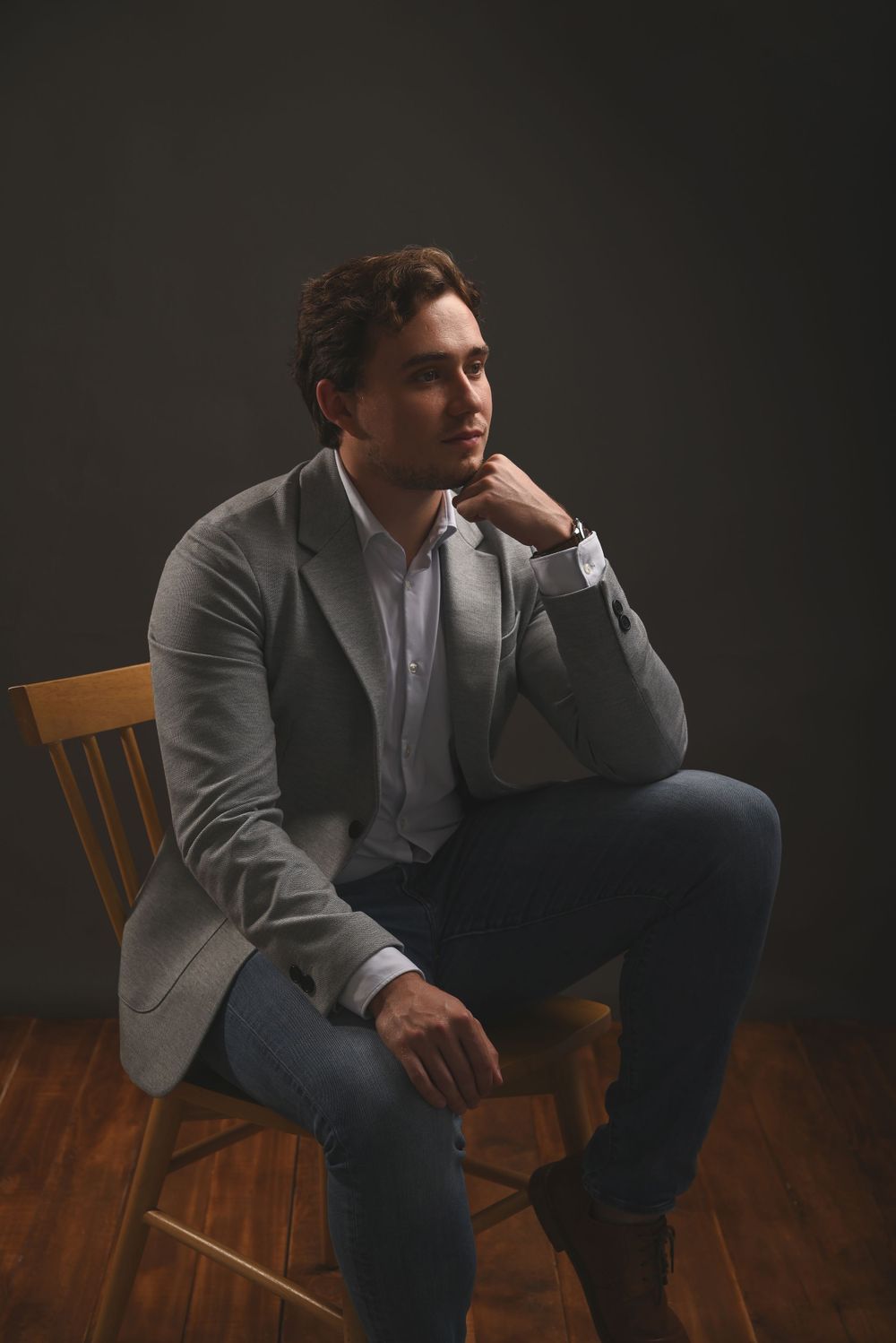 Professional portrait of someone in a gray blazer and jeans sitting thoughtfully on a wooden chair against dark background.