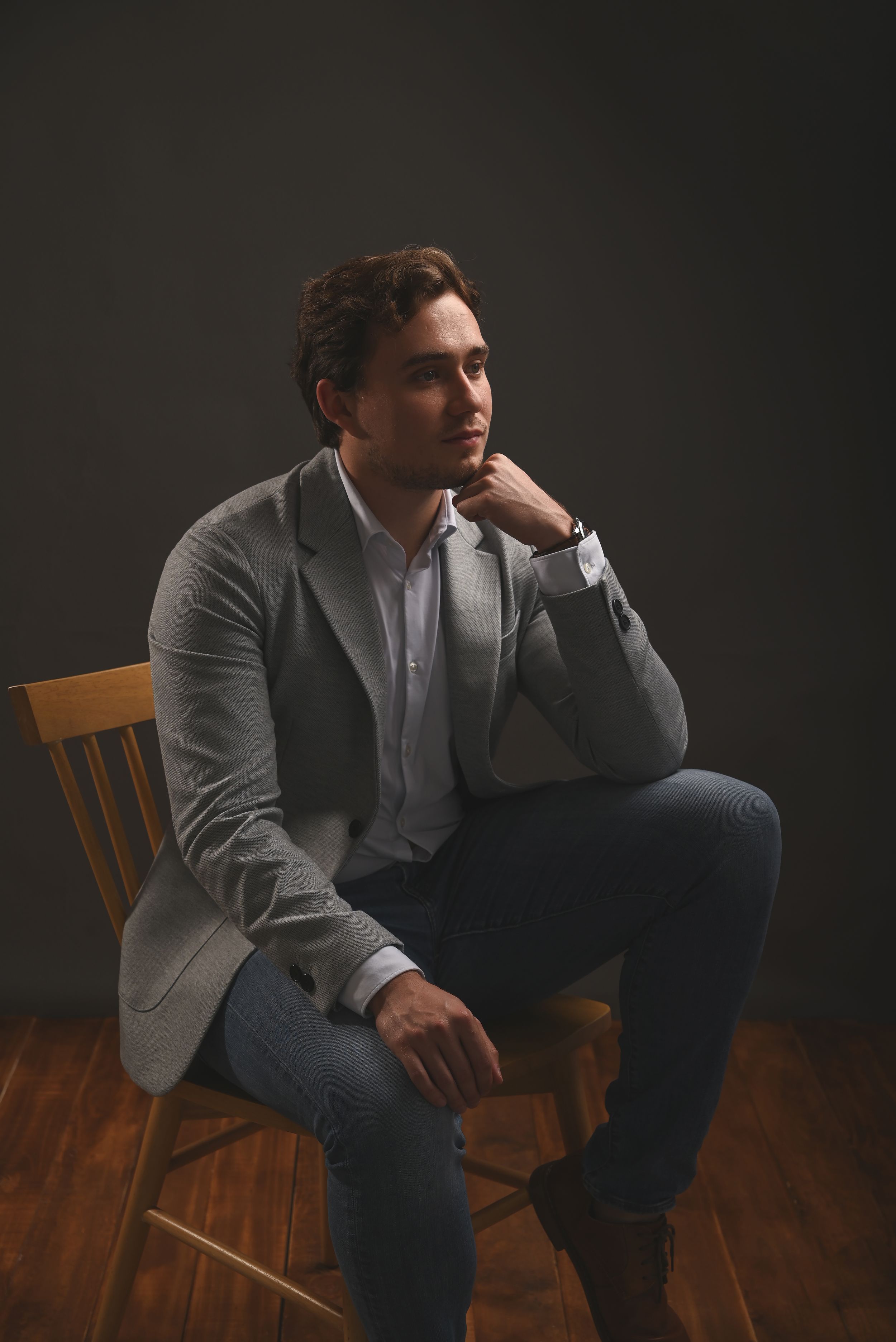 Professional portrait of someone in a gray blazer and jeans sitting thoughtfully on a wooden chair against dark background.