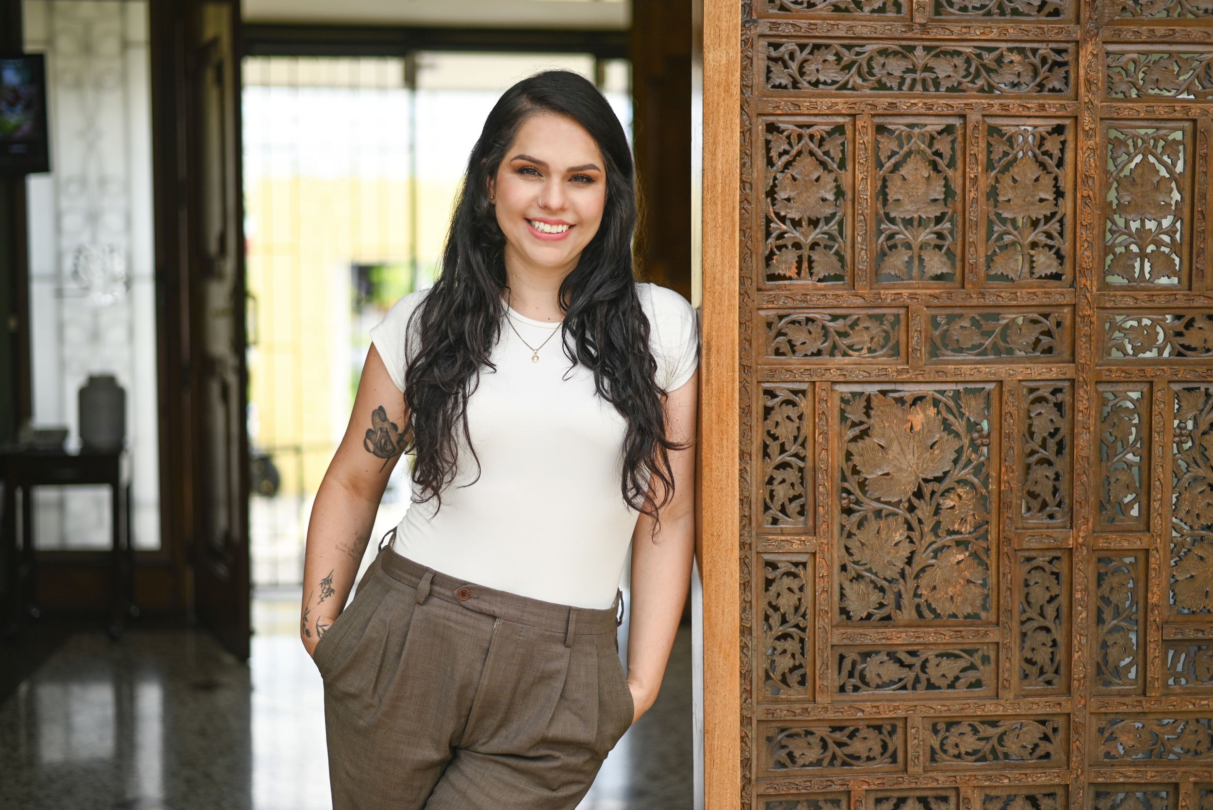 Someone in a white top and brown pants stands smiling next to an ornately carved wooden door in a bright corridor.