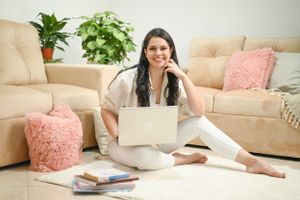 A person relaxes on a beige couch working on a laptop with pink pillows and houseplants decorating the living room.