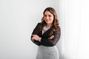 Professional studio photo of someone wearing a black sheer top and fitted gray skirt with statement necklace against white wall.