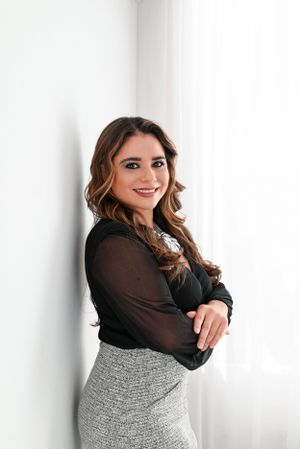 Professional portrait of a person in a black sheer top and textured grey pencil skirt standing against a white wall.